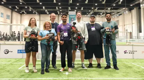 Seven men and women with some of them holding robots all standing in a line and facing the camera. They are standing on a small football pitch in a large arena with goals in the background.