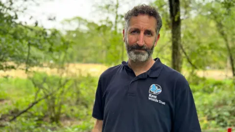 BBC / Fiona Irving A man with short curly brown hair and a greying beard looks at the camera. He is wearing a navy blue polo shirt with a butterfly logo and the words 'Kent Wildlife Trust' next to it. Behind him are green trees