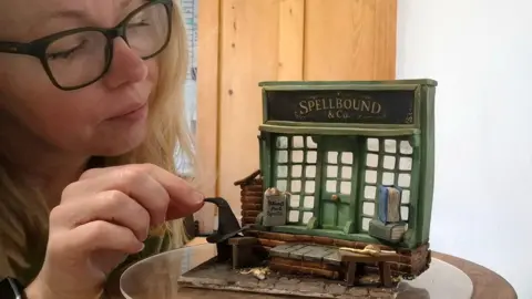 Niki Lowe adds a witch's hat to a minature shopfront made from cake and icing. The shop front has the sign Spellbound & Co 