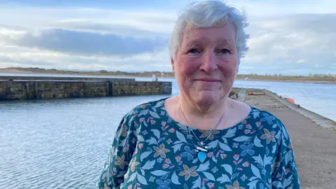 Jennifer Hall is a woman in her 60s with short grey hair who is wearing a dark blue top with a pattern of lighter blue flowers and leaves. She is standing on the harbour wall at Beadnell and behind her you can see the arc of the coast.
