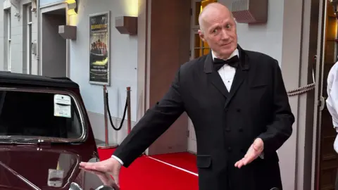 Adrian Edmondson wearing a formal black suit and bow tie stands on a red carpet outside a building with ornate architectural details. He's resting one hand on the Bergerac red triumph roadster and gesturing with the other. Behind is poster frames, exterior lights, a doorway, and velvet ropes lining the entrance area.
