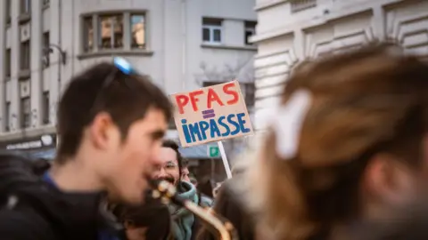 Hans Lucas_AFP/Getty Images A protest image, with blurred faces in the foreground, and in the background a man smiling with a beard and glasses is holding a sign that reads "PFAS = IMPASSE"