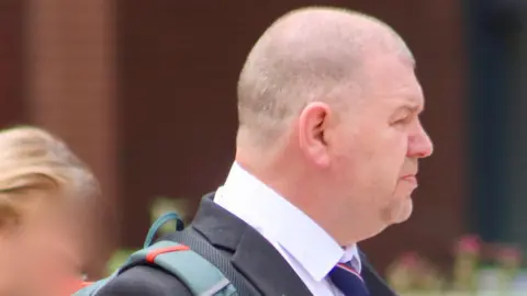 Lynda Roughley A side on view of Ian Ashworth, who has a shaven head and light stubble, walking away from court wearing a dark suit with a white shirt and blue and red striped tie.