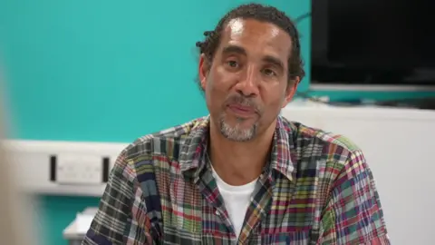 Paul Anderson sits at the table taking part in the discussion, a bright blue wall behind him. He has his hair tied back and wears a colourful plaid shirt
