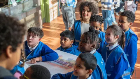 Liam Togher A group of children wearing blue uniform and hi-vis vests all hold a large white version of their book. They are stood in a hall and looking excitedly at something which is out of camera shot.