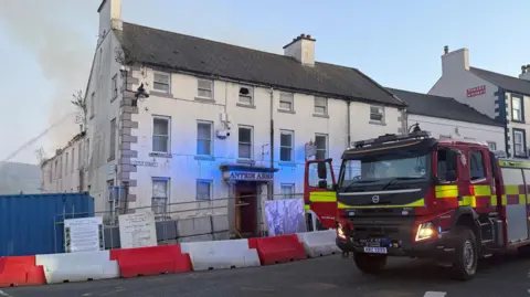 The Antrim Arms Hotel with a red fire engine in front of it. The building is white and Antrim Arms is written in red letters above the door. There are bollards and a fence in front of it. In the background, there is water being sprayed onto part of the building where white smoke is coming from.