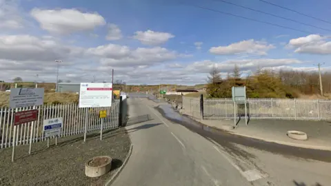 The entrance to Dunfermline Recycling Centre. There are several signs displaying information about the centre in front of a metal fence. A road runs through the middle leading into the centre. No buildings are visible.