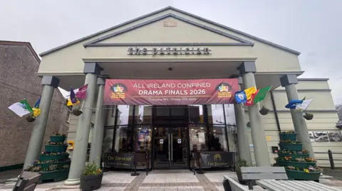 A building with an art deco facade. The building has pillars and a triangle front. County flags are attached to the pillars. Across the top is a banner that says 'All-Ireland confined drama finals'. There are glass doors into the hall, and large flower displays on either side of the doors. Across the front of the building it says The Patrician.