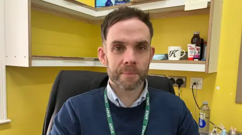 A man in an office. he is wearing a shirt and jumper