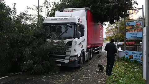 Getty Images A lorry with a tree on top of it and branches in front of it on a road