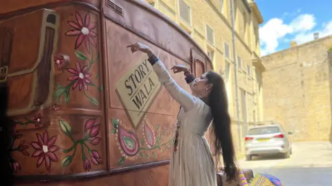 Grace Wood/BBC A woman with long brown hair and a white dress dances around a brown caravan
