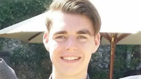 Headshot of Oliver McGowan, a young man with brown hair, standing outside and smiling