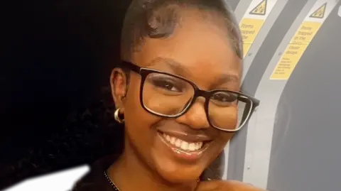 Elianne Andam is on a London Underground train. She is smiling and wearing glasses.