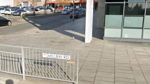 A google street view image of Millbay Road. A street sign is attached to a railing. There is a glass building to the right-hand side 