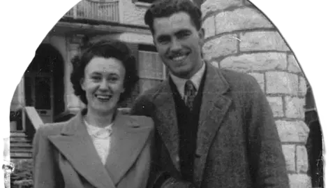 Steve Wilson A black and white cutout family photo of Pam and Charles, they are standing outside a brick building and both smiling at the camera. Pam is on the left, she has short dark hair fashioned into a blow dried perm, a padded shoulder coat with wide lapel collar and white blouse with a single string of pearls as a necklace. Charles is on the left, he has short dark hair and a moustache, wears a collared coact with a shirt and tie and v neck jumper.