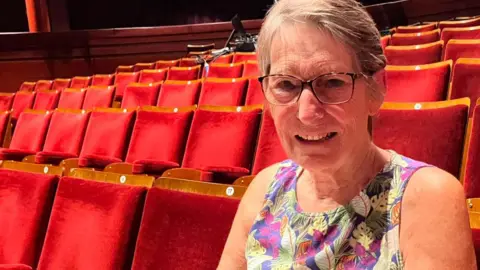 A woman sat in the theatre wearing a floral dress. She is smiling at the camera and is wearing black framed glasses with short hair. There are empty seats behind her.