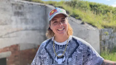 Rocca Holly-Nambi is standing inside the battery. They are wearing a gold chain around their neck and a cap with flower motif sewed onto the front of it.