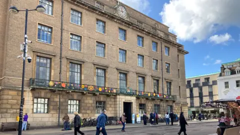 A four story Victorian style building with a balcony above the first floor. There is a clock at the top. People are walking on the pavement in front