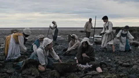 Contributed A still image from the Gossip film. A group of women sit and stand on a beach. They wear dresses, aprons and hats from the 16th Century.