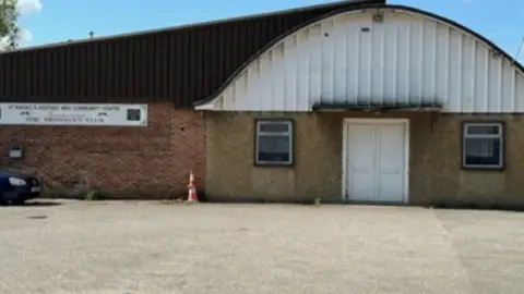 The centre, when it closed, with a dated facade 1940s facade of concrete and brick with white entrance door and a white arch-shaped roof above. A blurred sign on the left still has the former name St Marks and Hesters Way Community Centre on it 