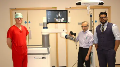 South Tees Hospitals NHS Foundation Trust Dr Simon Tizzard is wearing a blue medical hat, orange scrubs and glasses with Manju Prasad who has short hair, glasses, blue shirt and black trousers. Also in the picture is Dr Prasad Karpe, who has short black hair, black beard, blue shirt, blue waistcoat and trousers and dark stripy tie. They are standing with the robot in a hospital room.