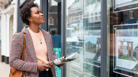 A woman looks in the window of an estate agent. She is wearing a smart jacket and peach jumper and holding a magazine. She is looking into the window and we see her face from the side