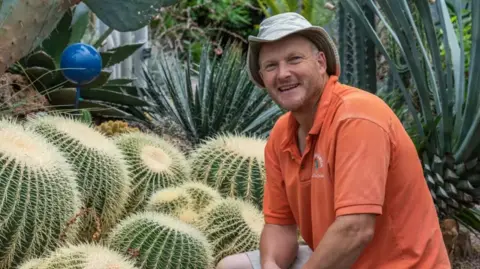 The World Garden Tom Hart Dyke wearing a hat and an orange top squatting down beside some cacti at The World Garden