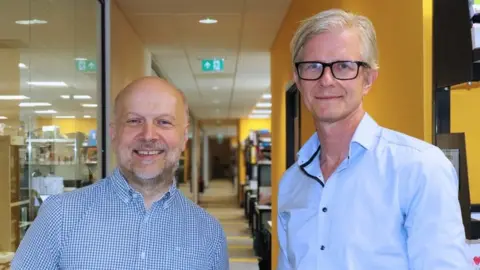 Cambridge Stem Cell Institute Bertie Gottgens and Matthias Zilbauer are standing side by side in a room that has yellow walls. Gottgens, on the left, has short grey hair and is wearing a blue and white shirt. Zilbauer on the right has short grey hair and is wearing black-framed glasses and a blue shirt.