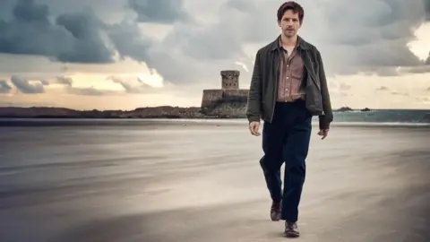 UKTV A man in smart casual clothes walks across a wide, flat sandy beach with a castle, sea and brooding sky behind him.