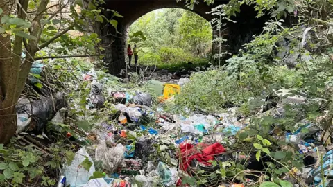 Network Rail Piles of rubbish mixed with foliage and overgrown trees littered all around the area. In the distance is a railway bridge with two volunteers in high-vis jackets standing underneath.