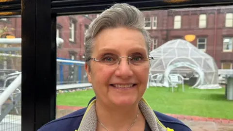 Andrea Jester has grey hair and glasses and is smiling. Grass and a building are in the background.