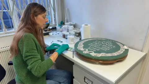 English Heritage A woman in a green jumper sits at a desk, working to restore the plaque which sits next to her 