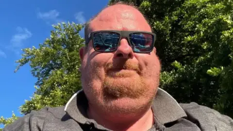 Richard Adams A head and shoulders selfie of a Richard Adams wearing black sunglasses and a grey shirt. There is blue sky in the background and a large tree.