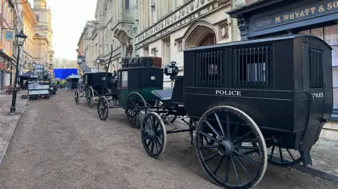 Part of Bristol city centre has been turned into an 1870s street for filming of Young Sherklock. Three old carriages are on the road, one is painted black with "Police" written on the side of it. A shopfront has been dressed to say M Whyatt & Sons in a traditional font.