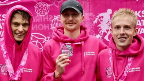 The Yorkshire Marathon The winner of the men's race shows off his medal while flanked by the runners who finished second and third. All three are wearing pink hoodies and smiling. They are standing in front of a pink Yorkshire Marathon banner. 