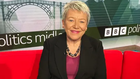 Rachel Taylor - a woman with short blonde hair and wearing a black suit jacket over a purple top, smiles as she sits on the Politics Midlands red sofa.