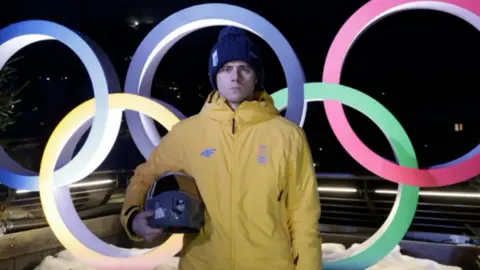 Vladyslav Heraskevych in a yellow jacket holds his helmet in from of the Olympic rings