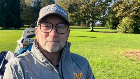 BBC Terry Kirby sits in a buggy in the sunshine on a green fairway at Ganstead Park Golf Club near Hull. He is wearing a light-grey jacket, matching "Titleist" cap and dark-rimmed glasses.