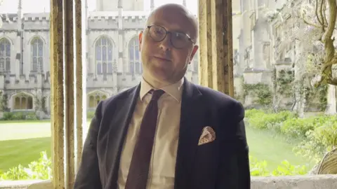Informator Choristarum Mark Williams has a bald head and glasses. He is wearing a blue blazer over a white shirt with a purple tie. Behind him is Magdalen College courtyard.