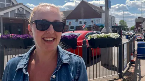 A woman smiling at the camera. She has blonde hair which is tied up in a ponytail and is wearing sunglasses. Behind her are railings and floral displays.
