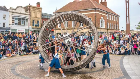 Large water wheel being pushed around a town centre in Derbyshire as part of a perfomance