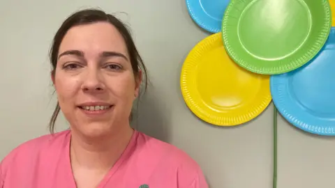 A woman with brown hair tied back, wearing pink scrubs.