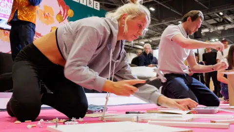 PA Media Hayley McAuley, who has blonde hair, a grey hoodie and black jogging pants is kneeling down and holding a hammer as she assembles a piece of flatpack furniture