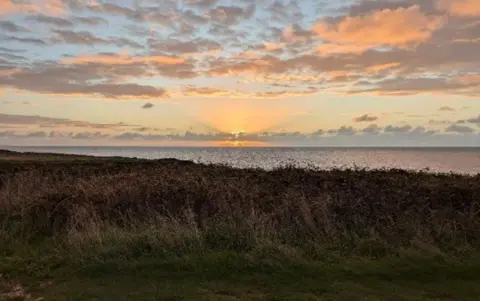 The sunrise, behind wispy clouds, casts a golden glow against a pale blue sky, above the sea in Weymouth Bay. in the foreground is brown vegetation and green grass.