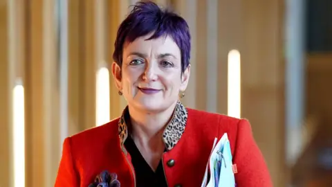 PA Media A smiling Angela Constance, who has short purple hair, walks along a wood paneled corridor in the Scottish Parliament. She is wearing a bright red blazer, with a leopard skin collar, and is carrying a pile of documents with her left arm.