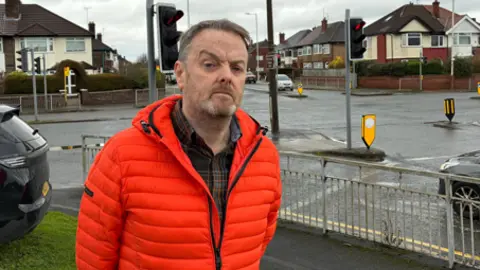 Man with brown hair and short beard stood at a road junction wearing a n orange jacket and looking at the camera.