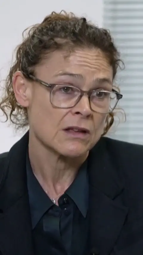 Jo Boydell looks slightly off camera. She wears brown plastic framed glasses and has light brown curly hair tied back. She wears a navy blue shirt and blazer.