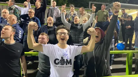 Warren Muggleton/BBC A group of Southend United fans cheering after their team won against Forest Green Rovers. Front and centre is a man looking straight at the camera with his arms in the air, wearing a white T-shirt. About 20 other fans are around him.