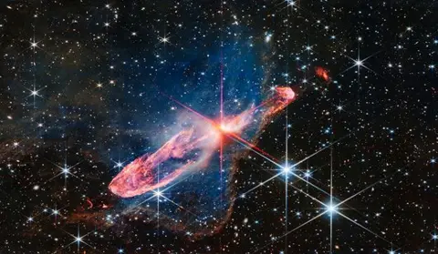 NASA A small bright red star in the centre in the process of forming as it illuminates a thick swirling ring of gas around it a deep red. In the background are a field of stars