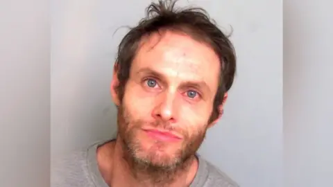 Essex Police A man with longish brown hair stares at the camera. He has blue eyes which look slightly red and a scruffy beard. He is wearing a grey top and stands against a grey background.